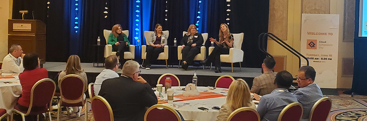 panel discussion of four women on a stage sitting in white wingback chairs in front of a large conference audience