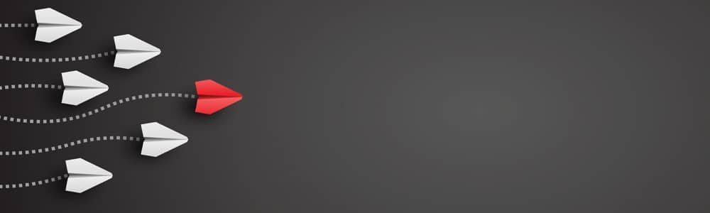 A red paper airplane leads several white paper airplanes in the same direction against a dark background.